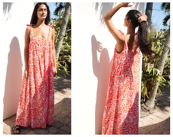 May include: A woman wearing a red and white floral print maxi dress with thin straps that tie at the shoulders. The dress has a square neckline and a loose, flowing fit.