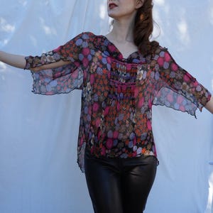 May include: A sheer, floral patterned blouse with a ruffled neckline and long sleeves. The blouse is a light pink, purple, and orange colour with a black background. The model is wearing black leather trousers.