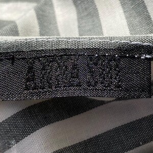 Pode incluir: Close-up de uma etiqueta preta com texto branco, costurada em tecido listrado cinza e branco. O texto da etiqueta &eacute; parcialmente vis&iacute;vel. O tecido tem uma textura tecida.