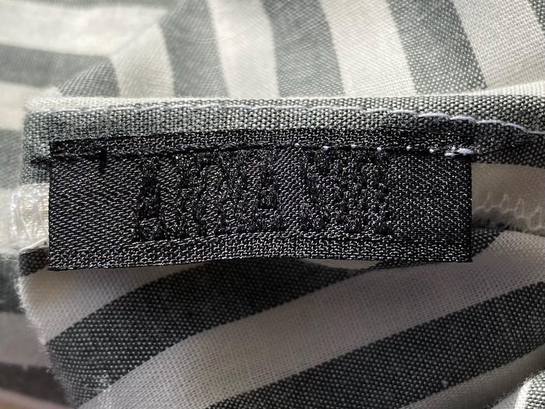 Pode incluir: Close-up de uma etiqueta preta com texto branco, costurada em tecido listrado cinza e branco. O texto da etiqueta &eacute; parcialmente vis&iacute;vel. O tecido tem uma textura tecida.