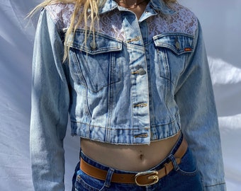 May include: A light blue denim jacket with a cropped hem and lace detailing on the shoulders. The jacket has a button closure and two front pockets.
