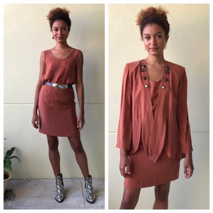 May include: A two-piece rust-colored outfit featuring a sleeveless dress and a matching jacket with decorative buttons. The jacket has a draped design and a slightly longer hemline than the dress.