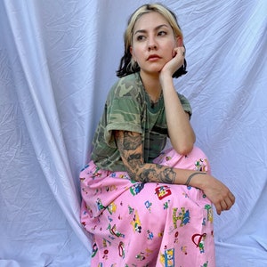 Op de afbeelding: Een camouflagegroen t-shirt en een roze pyjamabroek met tekenfilmprints. De broek is lichtroze en heeft verschillende tekenfilmfiguren. Het t-shirt heeft een camouflagepatroon.