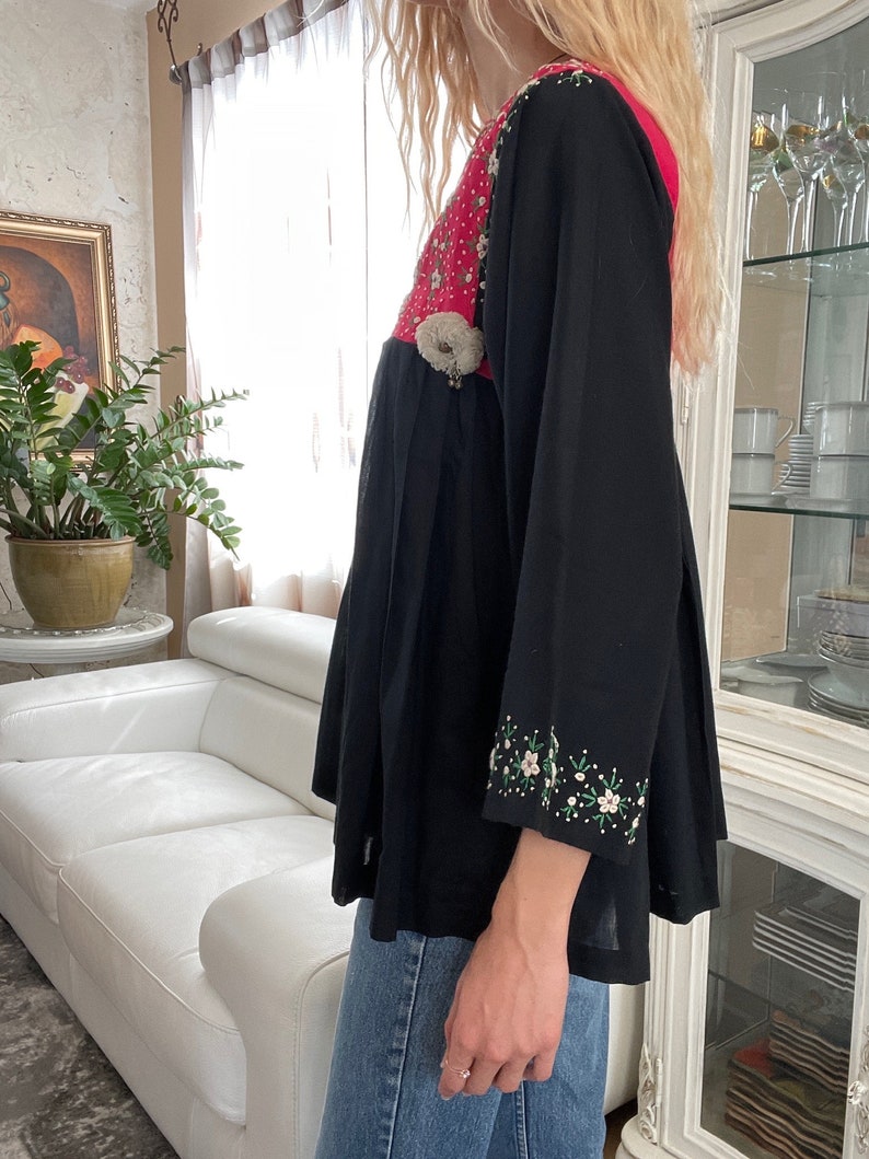 May include: Black tunic top with a pink and white floral embroidered bodice and sleeves. The top features a pleated skirt and a decorative embellishment at the waist. The sleeves are long and adorned with floral embroidery.