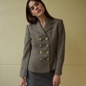 May include: A black and white patterned blazer with gold buttons. The blazer has a double-breasted closure and a collar. The model is wearing a gray skirt.