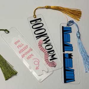 May include: Three clear acrylic bookmarks with different designs and tassels. The first bookmark has a pink worm with the text "BOOKWORM" in black. The second bookmark has the text "YOU FELL ASLEEP AROUND HERE" in pink. The third bookmark has a stack of blue books with the text "NEVER" in black.