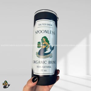 Pode incluir: Um copo preto e prateado de 591 ml com um rótulo branco com uma ilustração de sereia e o texto "SPOONLESS ORGANIC BRINE". O copo também tem o texto "SALTED SIREN" e "Chronically Ill".