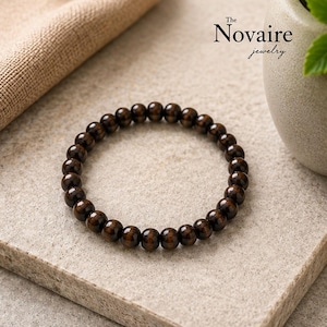 May include: A circular stretch bracelet composed of polished, dark brown wooden beads. The beads are uniform in size and strung together in a continuous loop. The text The Novaire jewelry is visible in the upper right corner of the image.