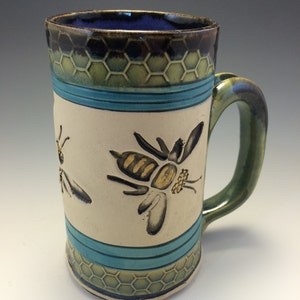 May include: A handcrafted ceramic mug with a cream-coloured body, adorned with two bee illustrations. The mug features a blue interior, a green handle, and a honeycomb pattern around the top and bottom. Turquoise stripes add a decorative touch.