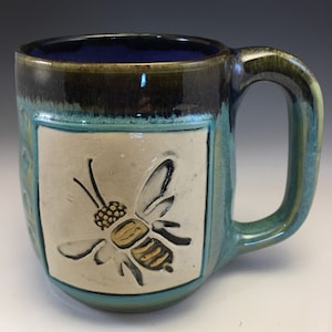 May include: Handmade ceramic mug with a bee design. The mug features a teal exterior with a rectangular cream-colored inset showcasing a detailed bee illustration. The interior is a deep blue, and the handle is a matching teal.