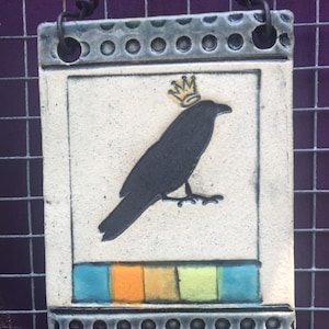 May include: Rectangular ceramic wall hanging featuring a black crow wearing a gold crown. The artwork has a cream-colored background with a border of blue-gray dots. The bottom has a row of colorful squares in blue, orange, and green. A black wire is attached for hanging.