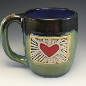 Puede incluir: Taza de cerámica hecha a mano con diseño de corazón. La taza presenta un corazón rojo en el centro de un diseño rectangular con rayos blancos, sobre un fondo verde y azul. El interior es de color azul oscuro.