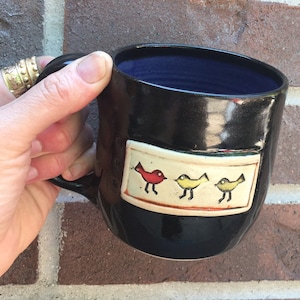 May include: Black ceramic mug with a dark blue interior. The mug features a rectangular design with three colorful bird illustrations. The handle is on the left side. The mug is held by a hand.