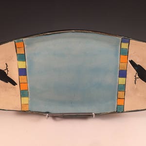 May include: Rectangular ceramic serving platter with a light blue center and tan ends. Each end features a black crow illustration. Vertical bands of orange, blue, and yellow squares border the blue center. The platter is supported by a metal stand.