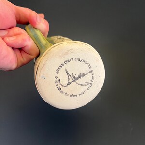May include: A handmade ceramic mug with a green handle and a cream-colored base. The bottom of the mug features the text "alissa clark clayworks" and a signature, along with the phrase "it's okay to play with your food."