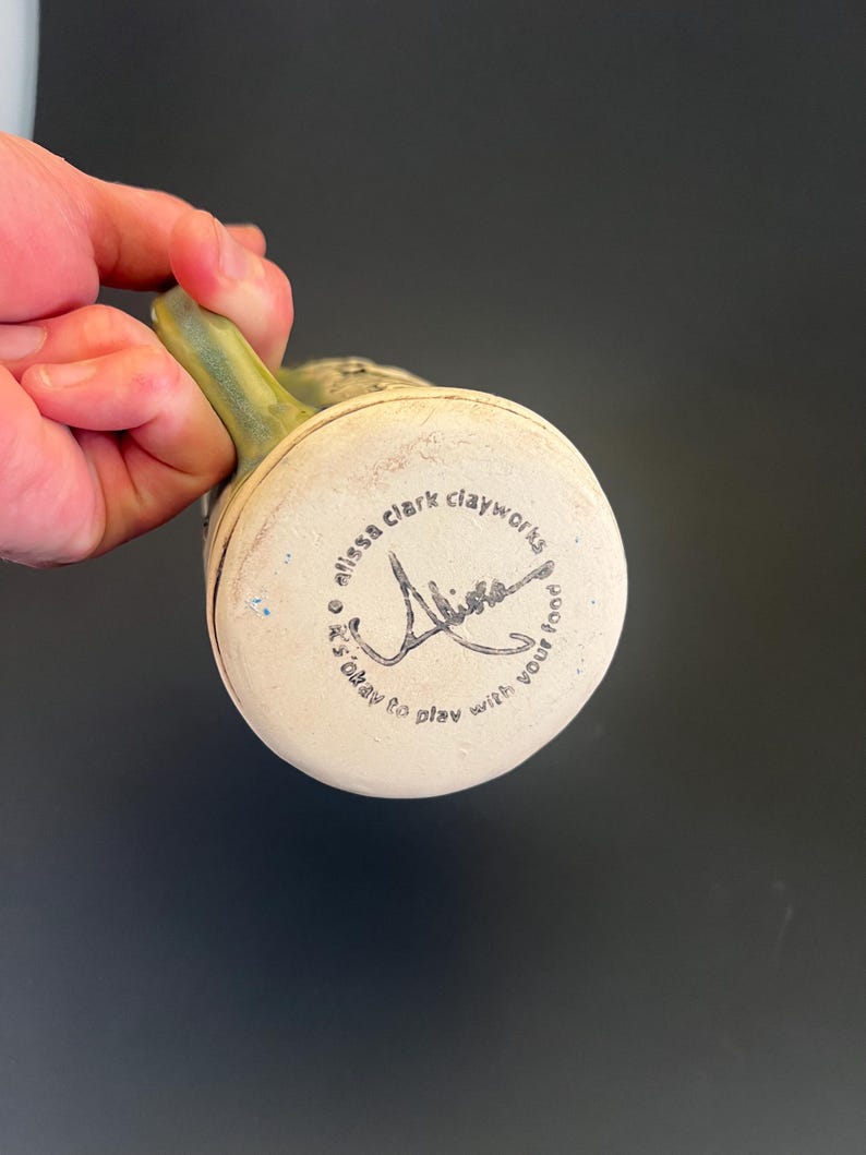 May include: A handmade ceramic mug with a green handle and a cream-colored base. The bottom of the mug features the text "alissa clark clayworks" and a signature, along with the phrase "it's okay to play with your food."