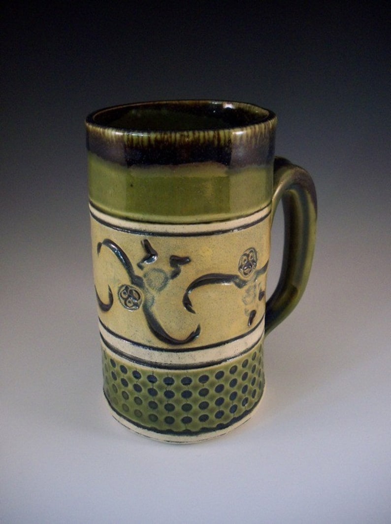 May include: Handmade ceramic mug with a green and tan color scheme. The mug features a dark brown rim, a green upper section, and a tan middle section with a decorative design. The bottom section has a dotted pattern.
