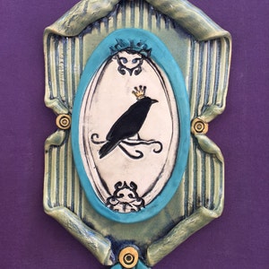 May include: A ceramic wall hanging featuring a black crow wearing a gold crown, perched on a branch within a teal oval frame. The frame is set against a striped green and blue background, with decorative floral accents and a purple backdrop.