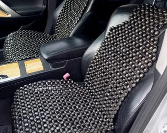 May include: A black beaded car seat cushion. The cushion is made of small, round beads that are woven together in a grid pattern. The cushion is placed on the driver's seat of a car.