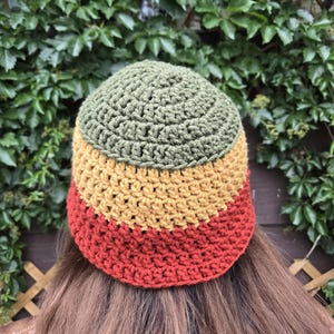 May include: A crocheted hat in a tri-color design. The top section is olive green, followed by a mustard yellow band, and a rust-colored brim. The hat is handmade and appears to be made of a soft yarn.