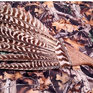 May include: A fan made of brown and white patterned feathers with a wooden handle. The feathers are arranged in a fanned-out shape, and the handle is a light brown color. The fan is set against a camouflage background.