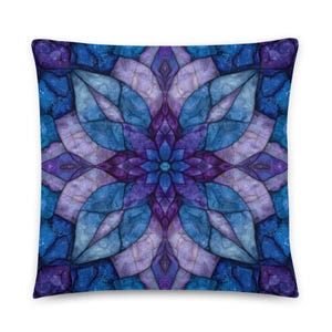 May include: Square throw pillow with a symmetrical floral design in shades of blue, purple, and lavender. The pattern resembles stained glass, with a central flower and radiating petal shapes. The pillow has a soft, textured appearance.