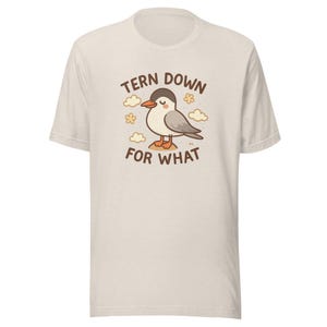 May include: Cream-colored t-shirt featuring a cartoon tern bird with the text "TERN DOWN FOR WHAT". The bird is gray, white, and brown, standing on a small island with clouds and flowers around it. A casual, fun graphic tee.