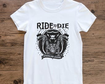 May include: White t-shirt featuring a black and white motorcycle graphic. The design includes the text "RIDE OR DIE" above a motorcycle image with a skull and the words "AMERICAN LEGEND" below.