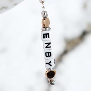 May include: A silver earring with a white bead charm that spells out "ENBY" in black letters. The earring has gold and silver accents.