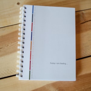 May include: A white spiral-bound journal with a vertical color-coded strip and the text "Today I am feeling...". The colors are green, red, and blue. The journal is on a wooden surface.