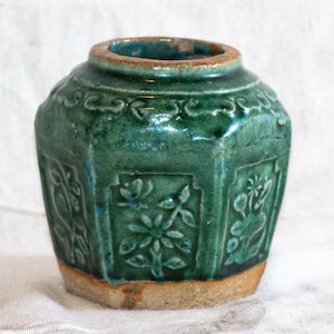 Op de afbeelding: Een kleine, antieke, groene keramische pot met een zeshoekige vorm en bloemreliëfpatronen. De pot heeft een bruine rand en bodem en staat tegen een witte achtergrond. De pot is een decoratief item.