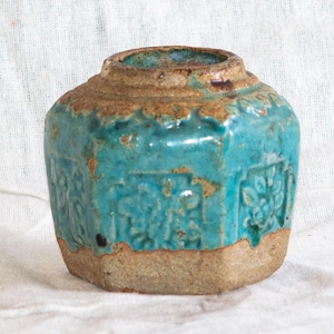 Puede incluir: Un pequeño jarrón de cerámica turquesa antiguo con un aspecto desgastado y texturizado. El jarrón presenta una base hexagonal y un detallado patrón floral. El borde tiene un acabado marrón envejecido, lo que añade encanto vintage.