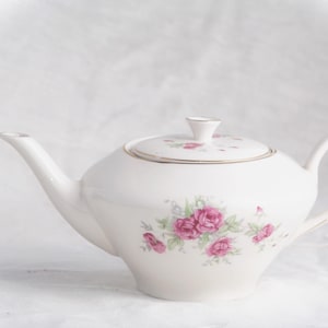 Può includere: Teiera in porcellana bianca con coperchio, con un motivo floreale di rose rosa e verdi. La teiera ha un beccuccio e un manico curvi, con finiture dorate. Un classico accessorio per il tè.