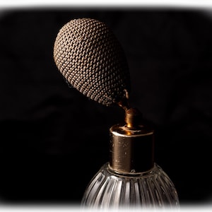 May include: An antique perfume bottle with a clear, ribbed glass base and a gold-colored metal collar. The atomizer is covered in a woven, dark brown material. The background is a dark, solid color.