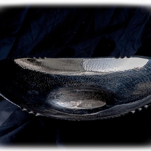 Puede incluir: Un cuenco de metal de forma ovalada con un acabado oscuro y reflectante, que presenta una textura martillada en toda su superficie. El cuenco tiene un diseño poco profundo y alargado con bordes ligeramente elevados y festoneados en el contorno.