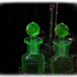 Può includere: Due bottiglie di profumo in vetro verde con tappi sfaccettati. Le bottiglie hanno una forma geometrica e sono esposte su uno sfondo scuro. Un supporto in metallo è visibile dietro le bottiglie.