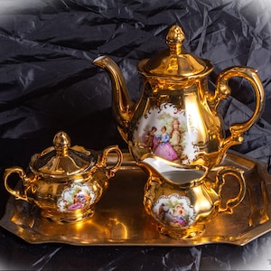 May include: A gold tea set comprising a teapot, creamer jug, sugar bowl, and tray. Each item has a painted scene of figures in historical dress. The set is presented on a reflective gold tray against a dark backdrop.
