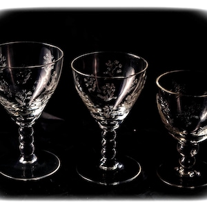 May include: Three clear glass goblets with floral etched designs. Each goblet has a round base, a twisted stem, and a flared bowl. The goblets are arranged in a row against a black background.