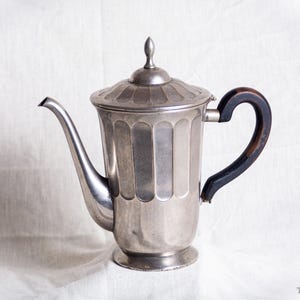 May include: A silver-colored coffee pot with a long spout and a dark wooden handle. The pot has a lid with a small finial and a decorative, vertical grooved pattern. The handle is black and curved. The pot sits on a white surface.
