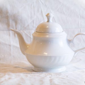 Könnte beinhalten: Eine weiße Keramik-Teekanne mit Deckel und gebogenem Griff. Die Teekanne hat einen Ausguss und steht auf einer weißen Oberfläche. Das Design ist schlicht und elegant, geeignet zum Servieren von Tee.