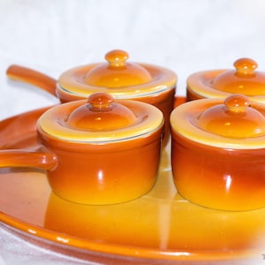 May include: Set of four orange ceramic mini casserole dishes with lids and handles, presented on a matching oval serving tray. The dishes have a glossy finish and a contrasting cream-colored rim.