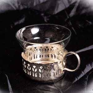 May include: A clear glass teacup sits in a decorative silver-colored metal holder with a handle. The holder has an ornate, openwork design. The teacup and holder are set against a dark, textured background.