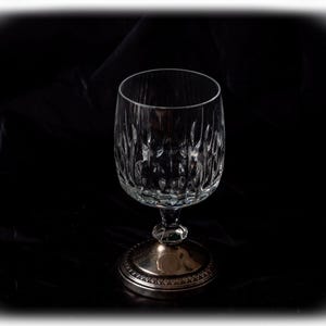May include: A clear crystal wine glass with a decorative pattern on the bowl and a silver-coloured base. The glass is empty and stands on a dark surface, creating a contrast. The stem has a faceted design.