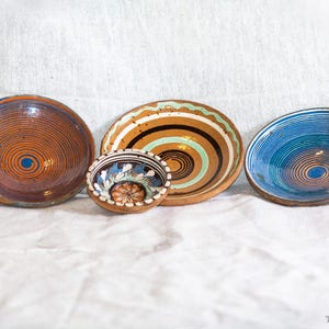 May include: Four handcrafted ceramic bowls with unique designs. The bowls feature spiral and concentric patterns in shades of brown, blue, and white. The smallest bowl has floral details. The bowls are arranged on a white cloth.