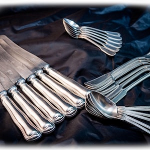 Puede incluir: Un conjunto de cubiertos de tono plateado que incluye cuatro cuchillos de mesa con mangos redondeados, un juego de cinco tenedores de cuatro dientes cada uno y dos grupos de cucharas. Los utensilios presentan un diseño clásico con superficies metálicas lisas.