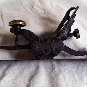 Op de afbeelding: Een antieke, donkere metalen tool met een complex ontwerp. Het heeft een cirkelvormige, messingkleurige knop, een gebogen handvat en diverse hendels en scharnieren. De tool rust op een textiel, off-white stof.