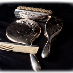 May include: A vintage silver vanity set, including a brush, hand mirror, comb, and a smaller brush. The set features floral engravings and is arranged on a dark surface. The brush bristles are light brown.