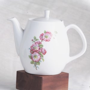 Könnte beinhalten: Weiße Keramik-Teekanne mit Deckel und Henkel, verziert mit einem Blumendesign aus rosa und roten Rosen mit grünen Blättern. Die Teekanne steht auf einem dunkelbraunen Holzblock.