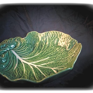 Peut inclure: Un bol en céramique verte en forme de grande feuille de chou avec une finition brillante et des nervures blanches proéminentes. Le bol présente un bord irrégulier et ondulé qui imite la texture naturelle d'une feuille. Le texte "Torch & Talon" est visible dans le coin.