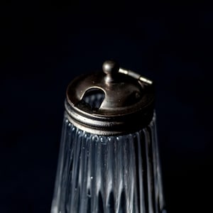 Peut inclure: Une saupoudreuse en verre transparent avec une texture côtelée verticale et un couvercle métallique argenté. Le couvercle comprend un petit bouton central et une ouverture circulaire pour verser, avec un mécanisme à charnière sur le côté.